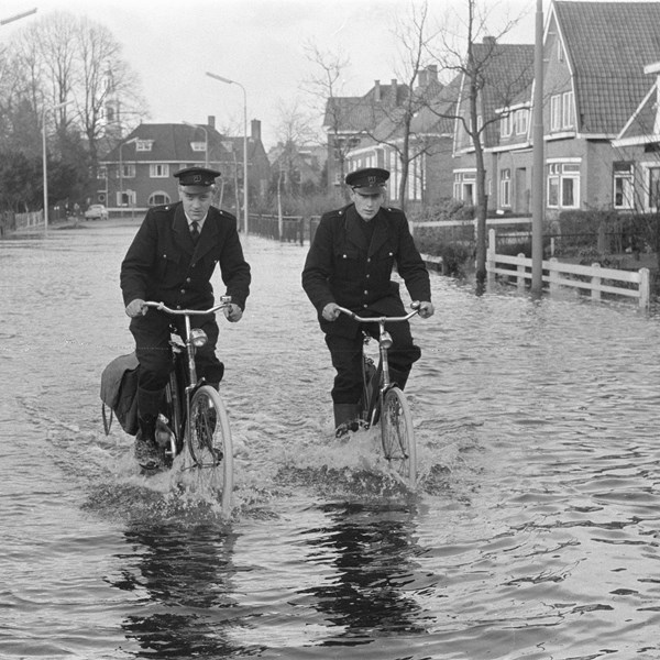 Twee postbodes van de PTT fietsen door hoogstaand water in Nijkerk in 1960