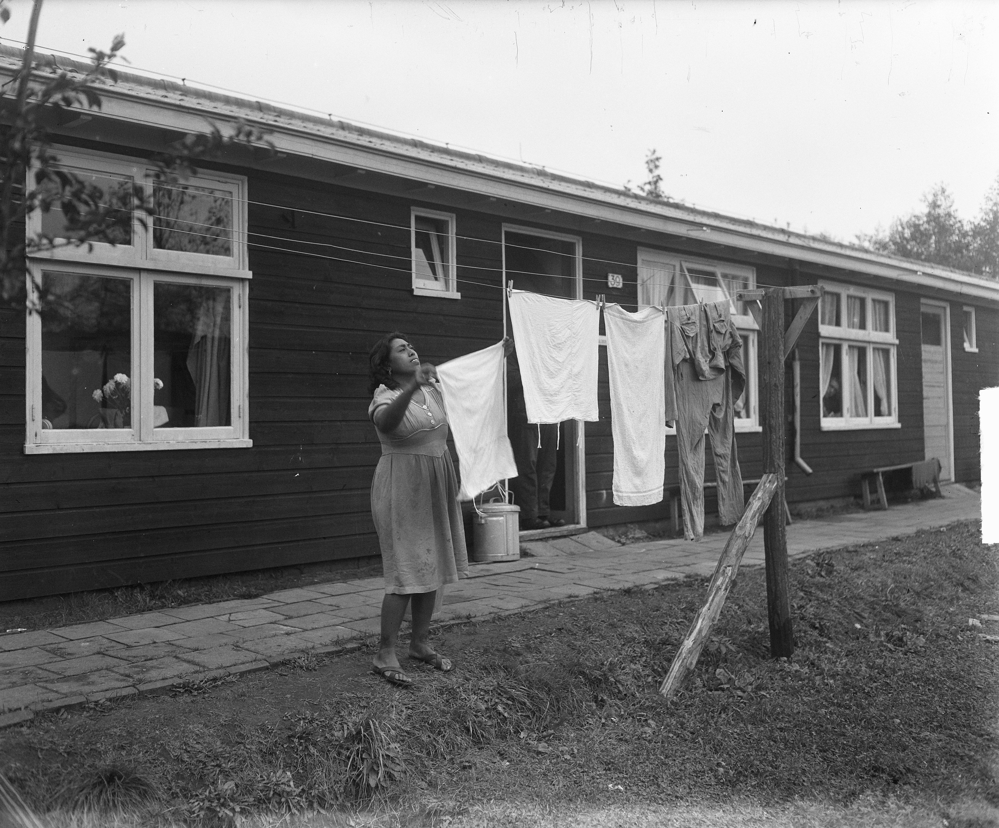 Foto van een vrouw die de was ophangt in Woonoord De Schattenberg in 1950; het voormalig kamp Westerbork. De eerste Indische Nederlanders arriveerden op 4 juli 1950 en verbleven daar tot 1951, toen het kamp werd vrijgemaakt voor Molukse bewoning. Bron: Nationaal Archief.