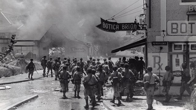 Foto van mariniers die een brandende wijk van Willemstad intrekken (hoek De Rouvilleweg) tijdens de opstand op Curaçao in 1969