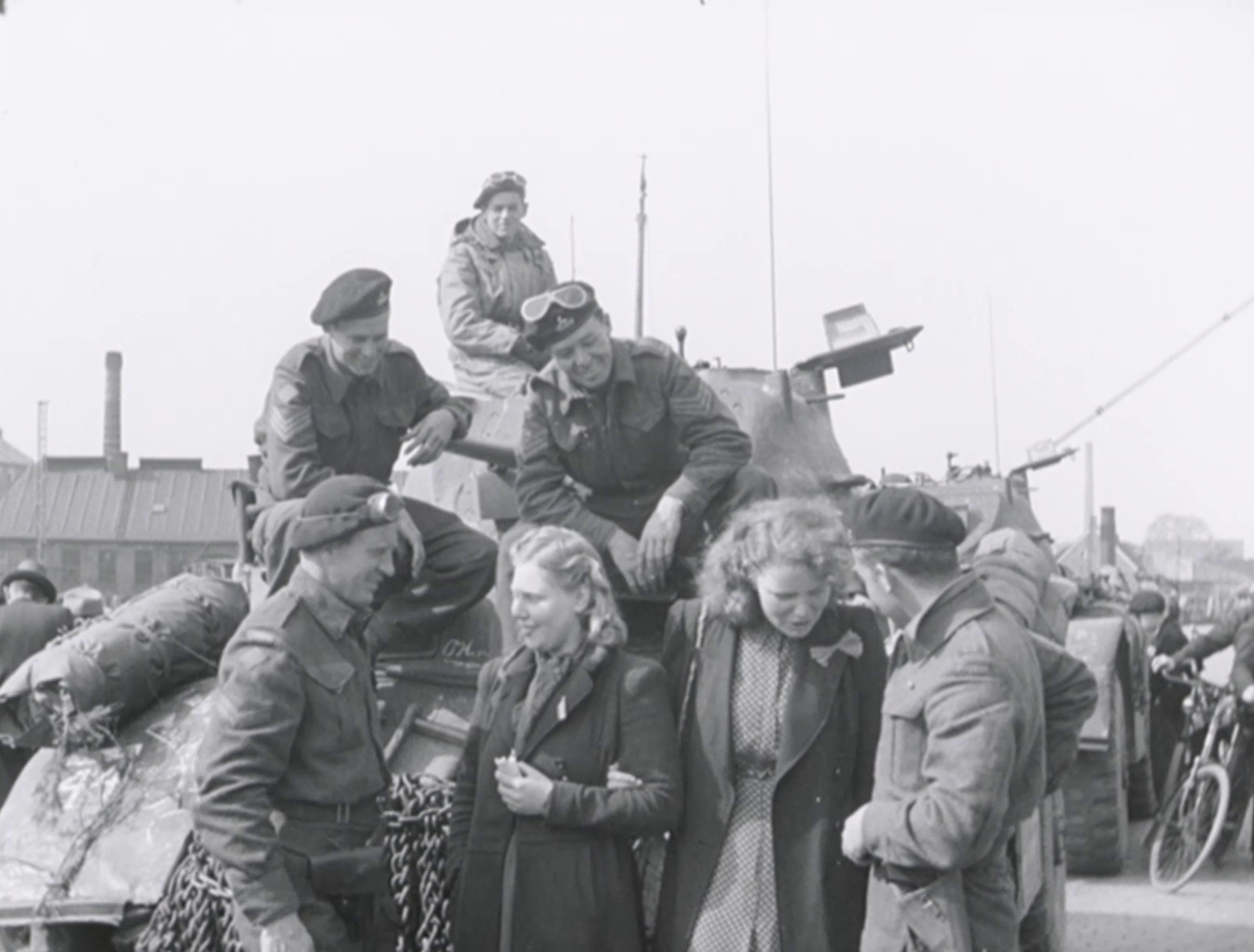 Zwart-wit-afbeelding van twee jonge blonde vrouwen die bij een tank staan, omringd door geallieerde soldaten te Groningen na de bevrijding.