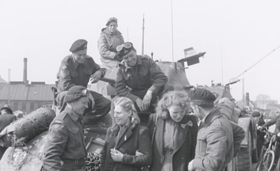 Zwart-wit-afbeelding van twee jonge blonde vrouwen die bij een tank staan, omringd door geallieerde soldaten te Groningen na de bevrijding.