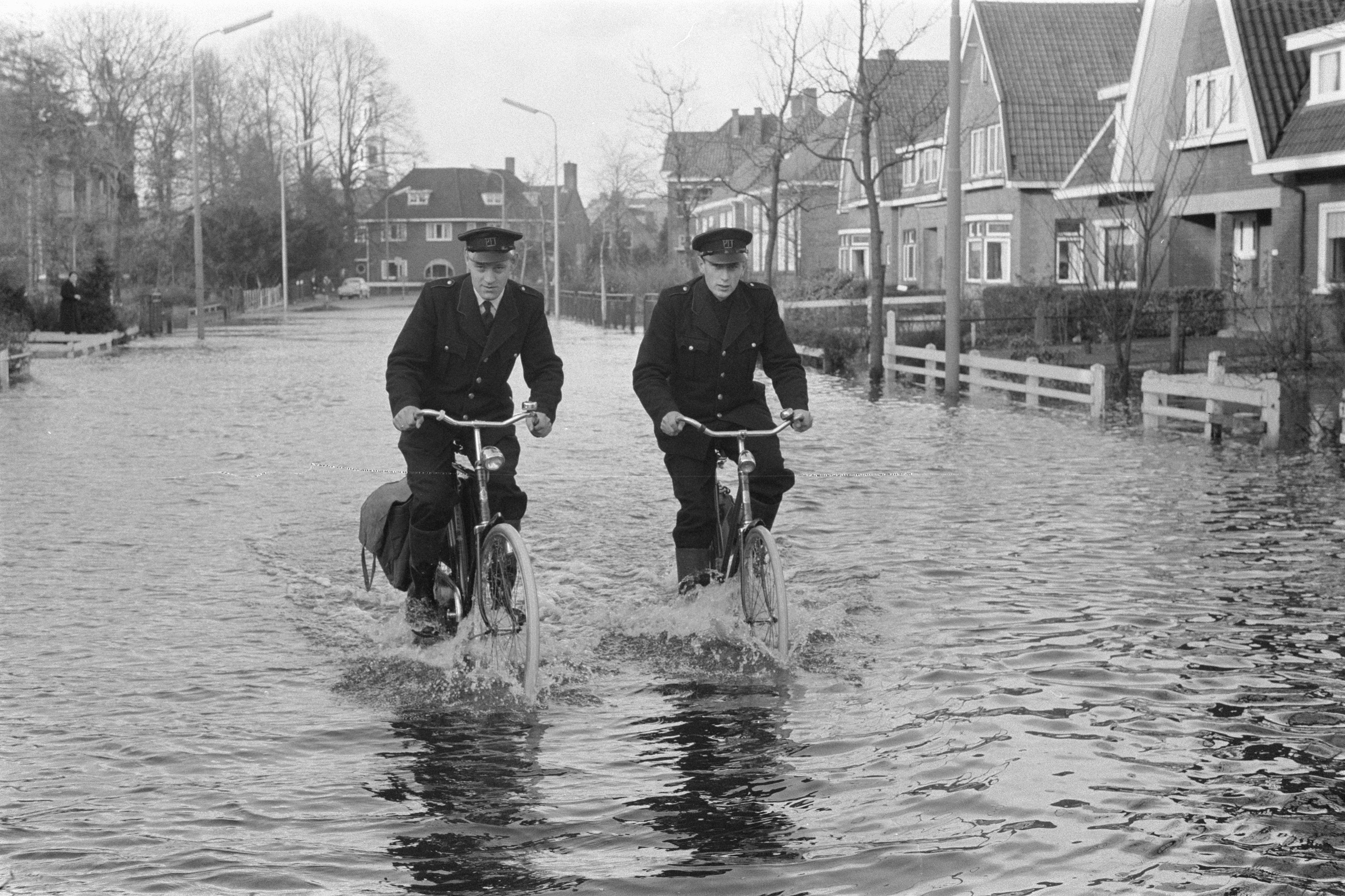 Twee postbodes van de PTT fietsen door hoogstaand water in Nijkerk in 1960